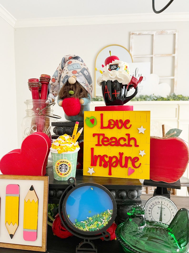 May include: A teacher-themed tabletop display with a gnome holding an apple, a coffee mug with an A+ on it, a yellow sign that says "Love Teach Inspire", a red apple, a globe, a green glass chicken, and a scale.