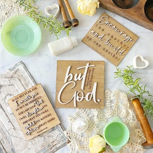 May include: An assortment of decorative wooden plaques with inspirational quotes, including "Encourage one another," and "but God." The image also features a green glass dish, a white milk bottle, and other decorative elements. The plaques are made of light-colored wood.