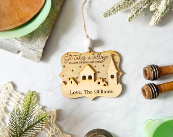 May include: A wooden ornament with a village design and the text "It takes a village." The ornament is light brown with a white bead and string for hanging. The text "Love, The Gilliams" is also included.
