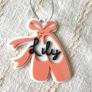 May include: A ballet slipper-shaped luggage tag, peach and white, with a bow detail and the name "Lily" in black script. It is attached to a silver wire loop. The tag is designed for luggage.