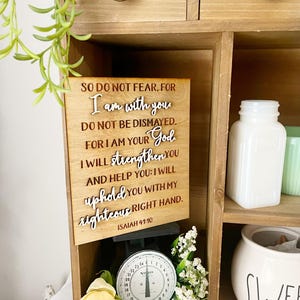 May include: Wooden sign with the text "SO DO NOT FEAR, FOR I am with you. DO NOT BE DISMAYED. FOR I AM YOUR God. I WILL strengthen You AND HELP YOU: I WILL uphold you WITH MY righteous RIGHT HAND. ISAIAH 41:10." The sign is displayed in a wooden cabinet with a vintage scale and decorative items.