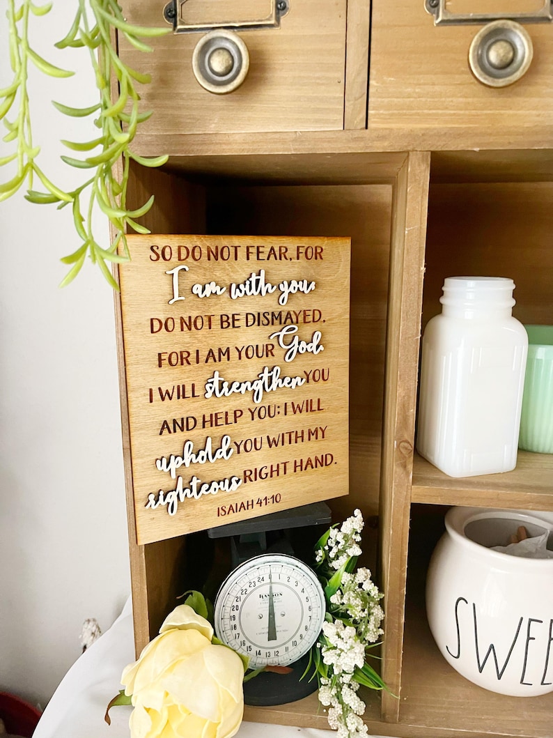 May include: Wooden sign with the text "SO DO NOT FEAR, FOR I am with you. DO NOT BE DISMAYED. FOR I AM YOUR God. I WILL strengthen You AND HELP YOU: I WILL uphold you WITH MY righteous RIGHT HAND. ISAIAH 41:10." The sign is displayed in a wooden cabinet with a vintage scale and decorative items.
