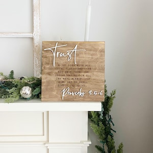Peut inclure: Panneau en bois avec le mot "Trust" en écriture blanche. Le panneau présente une citation inspirante de Proverbes 3:5-6. Le panneau est affiché sur une cheminée blanche avec de la verdure et une bougie.