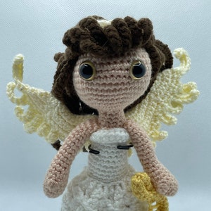 May include: A handmade crochet angel doll with brown hair, large black eyes, and cream-colored wings. The doll wears a white dress and blue shoes, and holds a yellow treble clef. The doll is standing on a black base.