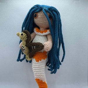 May include: A crocheted mermaid doll with long blue hair, holding a small crocheted seahorse. The mermaid is wearing a white and orange crocheted tail and a white crocheted top with an orange trim.