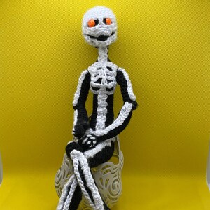 Puede incluir: Una muñeca de esqueleto de crochet con ojos naranjas y un diseño de esqueleto blanco y negro. La muñeca está sentada en una silla blanca con un diseño decorativo.