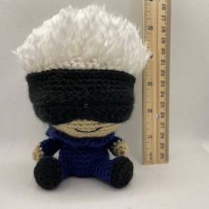 Puede incluir: Un juguete de peluche tejido a crochet de un personaje con cabello blanco, una máscara negra para los ojos y un mono azul. El juguete está sentado sobre una superficie blanca junto a una regla.