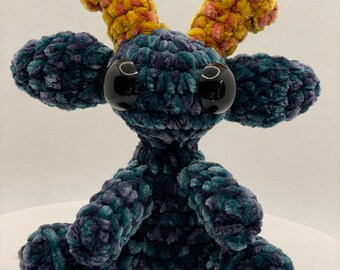 May include: A handmade plush creature with large, glossy black eyes and a textured, velvety body in shades of teal and purple. It has two yellow and pink antler-like appendages. The toy is sitting with its arms and legs outstretched.