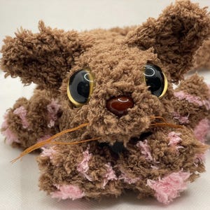 May include: A handmade brown plush toy bunny with large black eyes and gold rims. The bunny has pink accents on its paws and ears, with long whiskers. The toy is made of soft, textured material, and is designed to look like a rabbit.