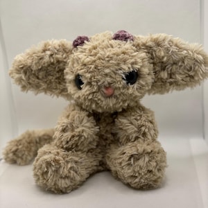 May include: A plush bunny toy in a seated position. The bunny is made of soft, light brown fur with large, black eyes and a pink nose. Two small, dark purple pom-poms are attached to the top of its head. The bunny has long, floppy ears.