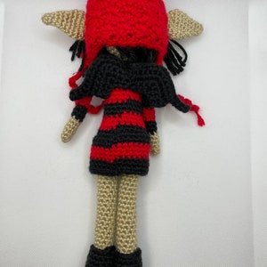 May include: A crocheted elf doll with black wings, a red and black striped dress, and a red crocheted hat. The doll has beige skin and black hair.