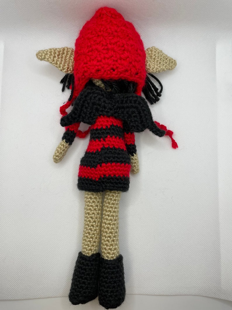 May include: A crocheted elf doll with black wings, a red and black striped dress, and a red crocheted hat. The doll has beige skin and black hair.