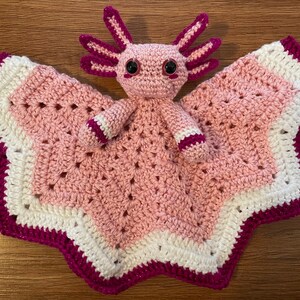 May include: A pink and white crocheted baby blanket with a pink axolotl amigurumi toy attached. The blanket has a scalloped edge with a pink trim.