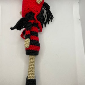 May include: A crocheted doll with red and black striped clothing, a red hat with horns, and black boots. The doll has long black hair and wings.