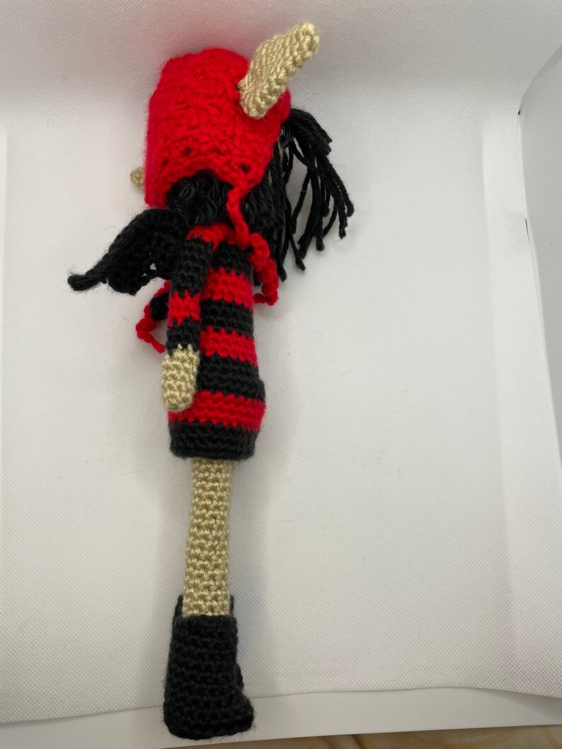 May include: A crocheted doll with red and black striped clothing, a red hat with horns, and black boots. The doll has long black hair and wings.