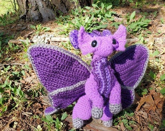 Puede incluir: Un dragón de crochet morado con detalles grises y alas grandes. El dragón está sentado en un lecho de agujas de pino y hierba verde.