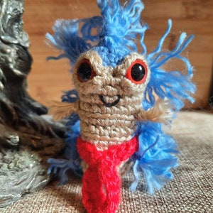 May include: A blue and brown crocheted toy with red yarn for a scarf. The toy has large, red eyes and a smiling mouth.