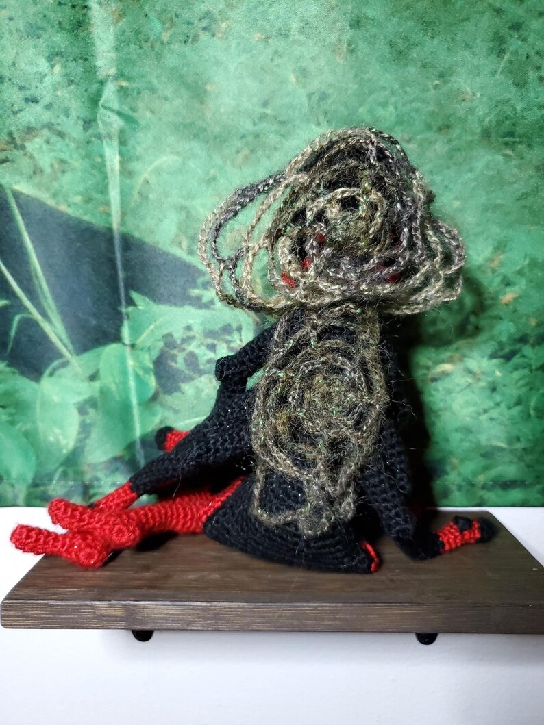 May include: A crocheted doll with black and green yarn for the body and hair, and red yarn for the legs. The doll is sitting on a wooden shelf.