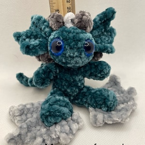 May include: Teal blue crocheted dragon plush toy with gray and white accents. The dragon has large blue eyes and is approximately 4 inches tall. Message for color selection/availability.