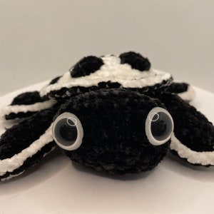 May include: A black and white crocheted turtle plush toy with large black eyes. The turtle has a white shell with black spots and a black body.