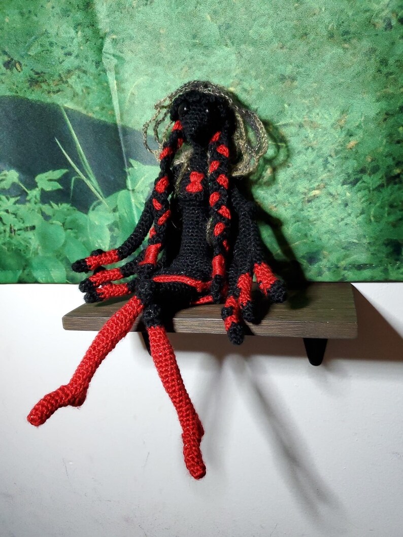 May include: A crocheted doll with black and red yarn. The doll has long, red yarn hair and is sitting on a wooden shelf.