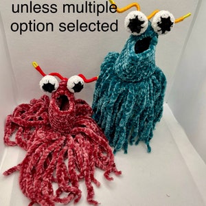 May include: Two crocheted plush toys with googly eyes and long, stringy hair. The toy on the left is pink and the toy on the right is teal. The text "Sold Individually unless multiple option selected" is visible at the top of the image.