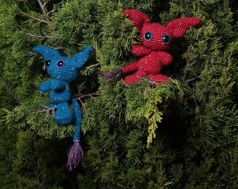 May include: Two handmade crochet creatures with large eyes and pointed ears are nestled in evergreen branches. One is teal with a purple tail, the other is red. Both have large, blue eyes and are posed as if perched in the tree.