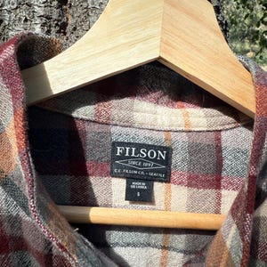 Pode incluir: Uma camisa de flanela xadrez vermelha, marrom e cinza com uma etiqueta Filson. A etiqueta diz "Filson Since 1897 C.C. Filson Co. - Seattle" e "Made in Sri Lanka S".