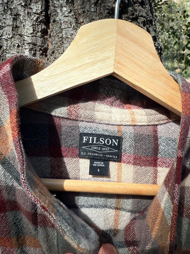 Pode incluir: Uma camisa de flanela xadrez vermelha, marrom e cinza com uma etiqueta Filson. A etiqueta diz "Filson Since 1897 C.C. Filson Co. - Seattle" e "Made in Sri Lanka S".