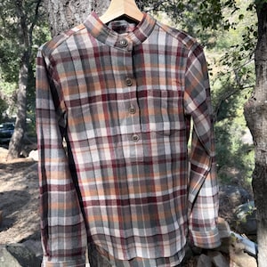 Pode incluir: Uma camisa de manga comprida xadrez com colarinho mandarim e fecho de bot&otilde;es. A camisa &eacute; xadrez marrom, bege e vermelho.