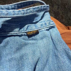 K&ouml;nnte beinhalten: Nahaufnahme eines hellblauen Jeanshemdes mit einem braunen Leder-Wrangler-Etikett.