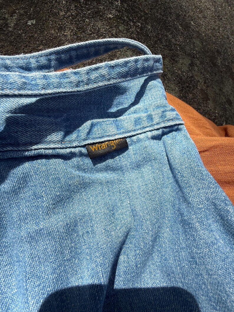 K&ouml;nnte beinhalten: Nahaufnahme eines hellblauen Jeanshemdes mit einem braunen Leder-Wrangler-Etikett.