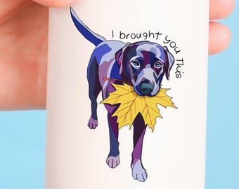 Pode incluir: Um copo de viagem de aço inoxidável branco com tampa, apresentando um gráfico colorido de um cão de pelagem escura segurando uma folha de outono amarela na boca, acompanhado pelo texto "I brought you this" em uma fonte cursiva preta.