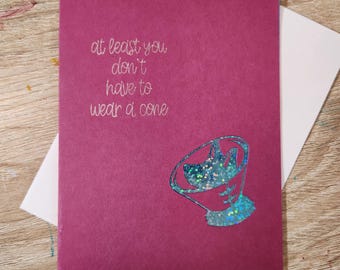 May include: A magenta greeting card with the text "at least you don't have to wear a cone" in silver script. A glittery blue cone shape is on the bottom right. The card is on top of a white envelope.