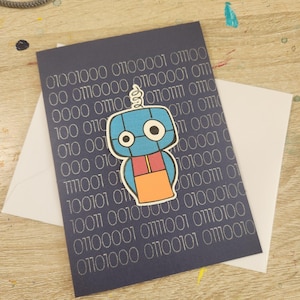 May include: A navy blue greeting card with a silver binary code pattern and a cartoon robot sticker. The robot is blue, orange, and red. A white envelope is behind the card.