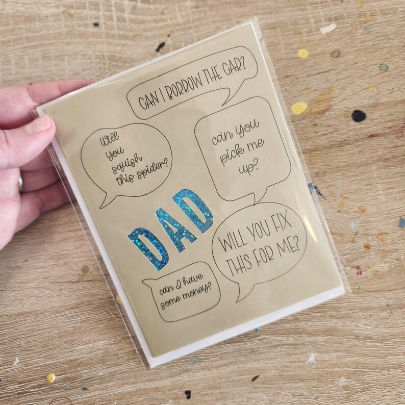 Pode incluir: Um cart&atilde;o de felicita&ccedil;&otilde;es bege com a palavra "DAD" em glitter azul e bal&otilde;es de fala com frases como "Posso pedir o carro emprestado?" e "Voc&ecirc; conserta isso para mim?"