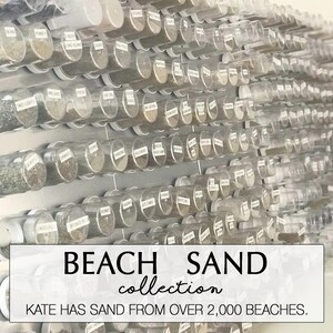 May include: A collection of beach sand samples displayed in clear tubes, each labeled with a beach name. The tubes are arranged in rows on a wall. The text "BEACH SAND collection" and "KATE HAS SAND FROM OVER 2,000 BEACHES" are visible.