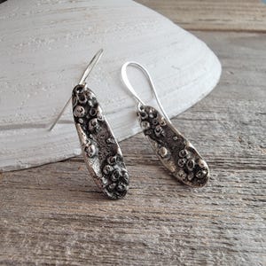 May include: A pair of silver-toned drop earrings. Each earring has a textured, oblong design with a series of small, raised circular details. The earrings hang from a simple hook. The background includes a seashell and a wooden surface.