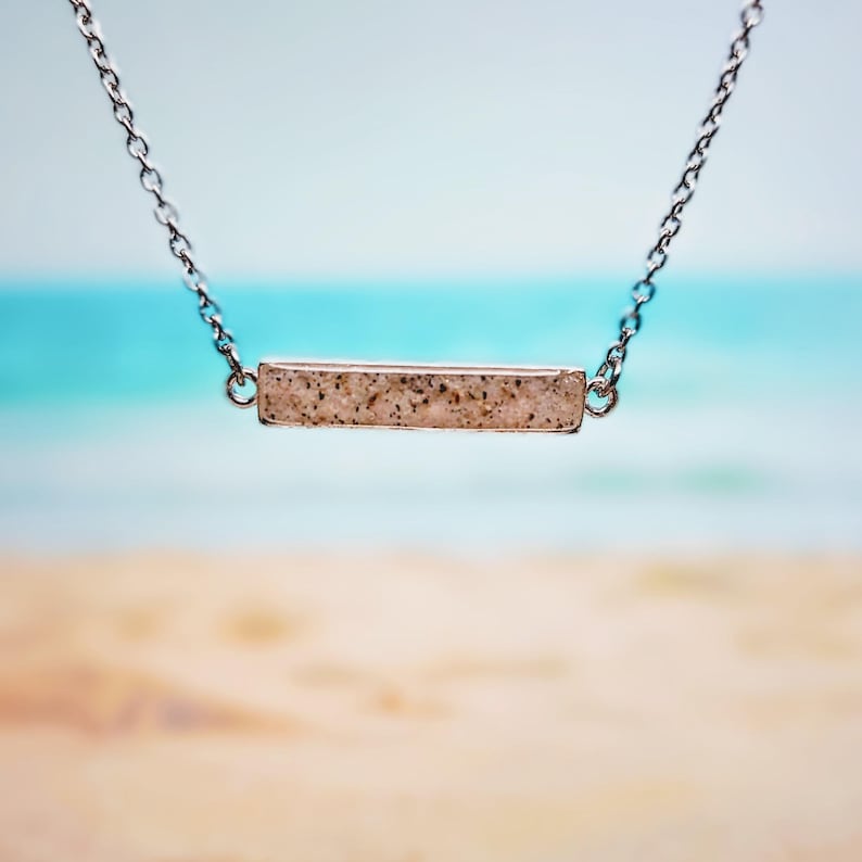 May include: A silver necklace with a rectangular pendant. The pendant has a speckled, light-colored appearance, possibly made of stone or resin. The background is a blurred beach scene with blue water and sand.