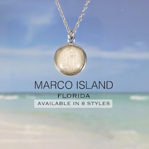 May include: A silver-toned necklace with a round pendant. The pendant contains a light-colored, textured material, possibly sand. The text "MARCO ISLAND FLORIDA" is below the necklace, with "AVAILABLE IN 8 STYLES" below that.
