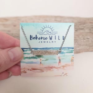 May include: A silver-toned necklace with a rectangular pendant filled with sand-like material. The necklace is displayed on a card with the text "Boheme WILD JEWELRY" and a beach scene illustration. The necklace is held by a hand.