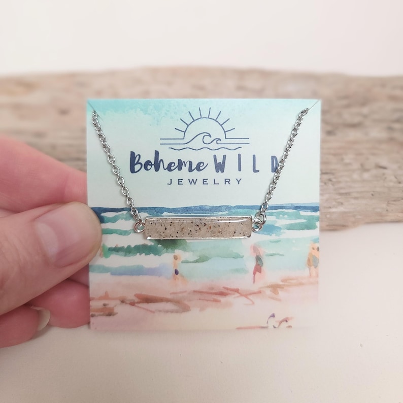 May include: A silver-toned necklace with a rectangular pendant filled with sand-like material. The necklace is displayed on a card with the text "Boheme WILD JEWELRY" and a beach scene illustration. The necklace is held by a hand.