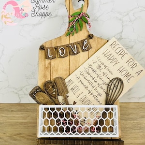 May include: Wooden kitchen decor with a cutting board that says "LOVE" and a recipe card that says "RECIPE FOR A HAPPY HOME" with a list of ingredients. The cutting board has a heart-shaped handle and a small wire basket.