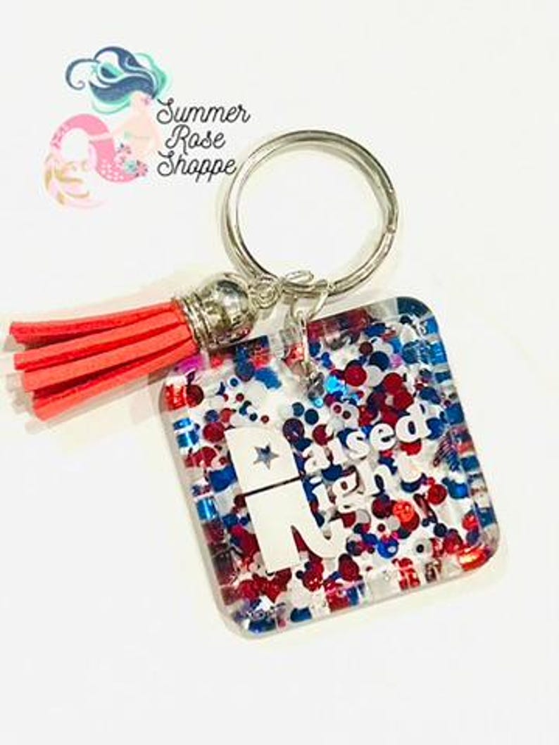 May include: A square keychain with a silver ring and a red tassel. The keychain has a clear background filled with red, white, and blue glitter and the text "Raised Right" with a star. The keychain is from Summer Rose Shoppe.