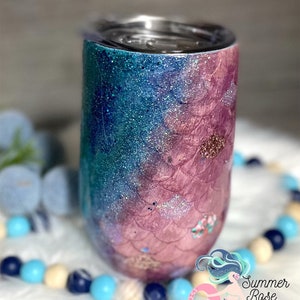 May include: A glittery tumbler with a mermaid scale design. The tumbler transitions from teal to pink with silver glitter accents. The design includes a mermaid scale pattern and small floral details.