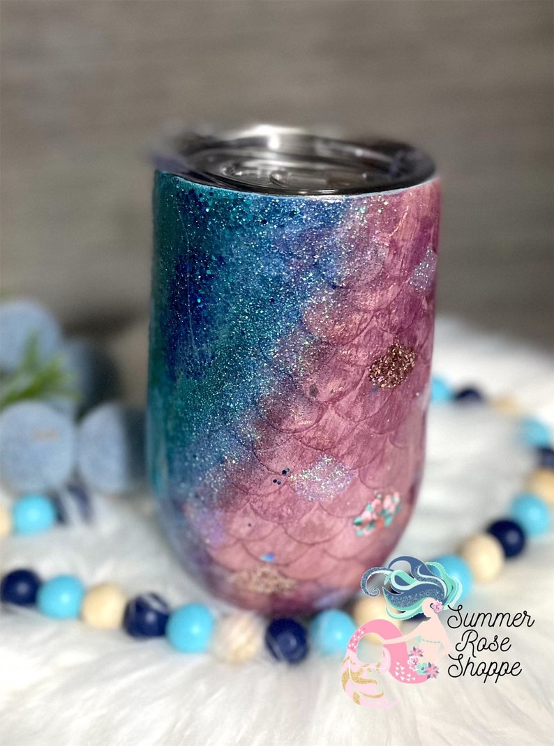 May include: A glittery tumbler with a mermaid scale design. The tumbler transitions from teal to pink with silver glitter accents. The design includes a mermaid scale pattern and small floral details.