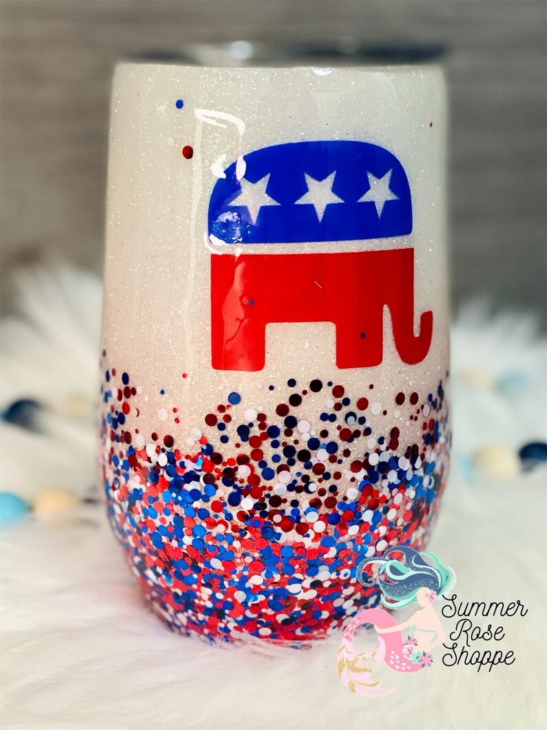 May include: A stemless wine tumbler with a glittery white finish. The tumbler features a red, white, and blue Republican Party elephant logo. The bottom of the tumbler is covered in red, white, and blue glitter.