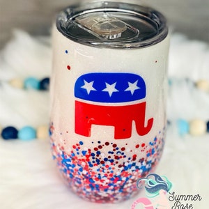 May include: A white glitter tumbler with a silver lid. The tumbler features a red, white, and blue elephant logo. The bottom of the tumbler is covered in red, white, and blue glitter. The tumbler is a stemless wine glass shape.
