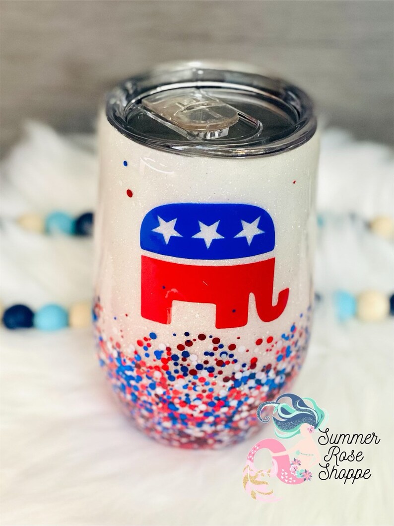 May include: A white glitter tumbler with a silver lid. The tumbler features a red, white, and blue elephant logo. The bottom of the tumbler is covered in red, white, and blue glitter. The tumbler is a stemless wine glass shape.