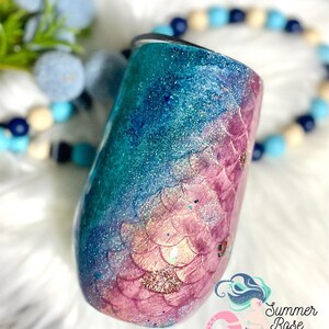 May include: A stemless wine tumbler with a mermaid scale design. The tumbler features a gradient of teal and pink glitter, with a scale pattern on the lower half. The tumbler has a silver rim and a black lid. The Summer Rose Shoppe logo is visible.